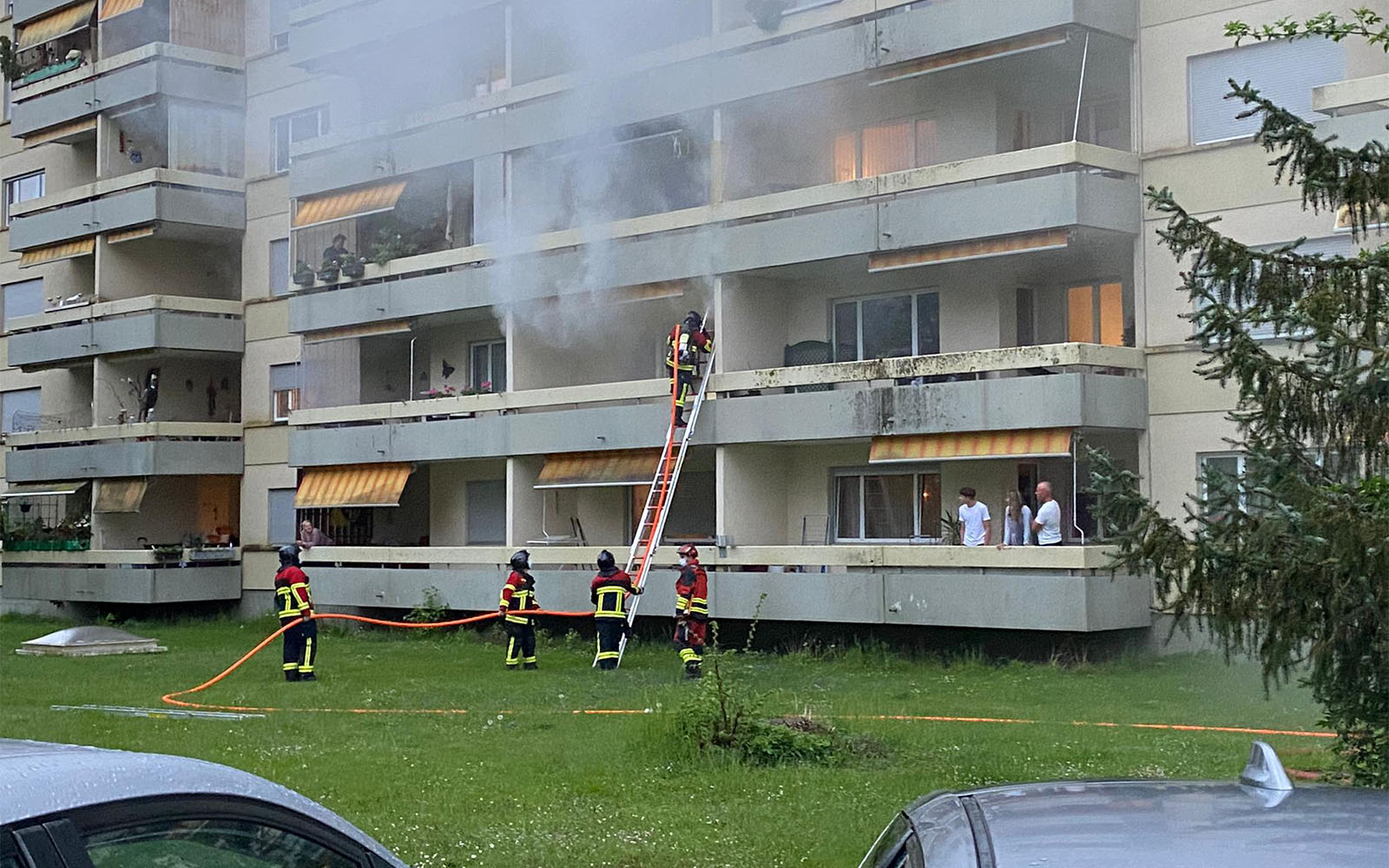 Fünf Feuerwehrleute löschen mit einem Strahlrohr ein Feuer in einem Wohnblock. Nachbarn stehen auf den Balkonen und sehen zu.